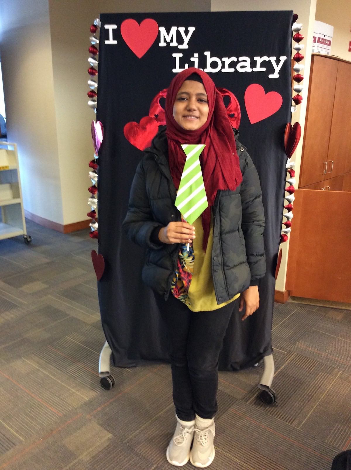 A student wearing a red hijab and puffy jacket poses in front of the I love my library backdrop holding up a prop necktie on a stick and smiling